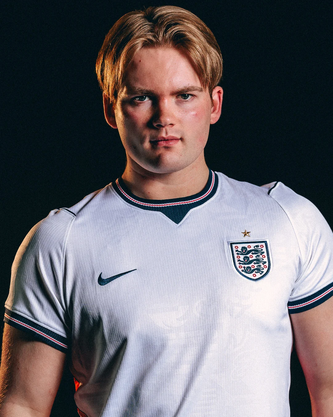 Studio portrait of a young blonde man wearing an England football jersey against a black background.