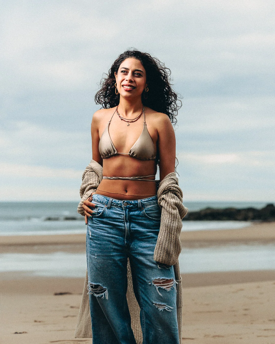 A woman with curly hair posing on a beach wearing a gold bikini top and ripped blue jeans.