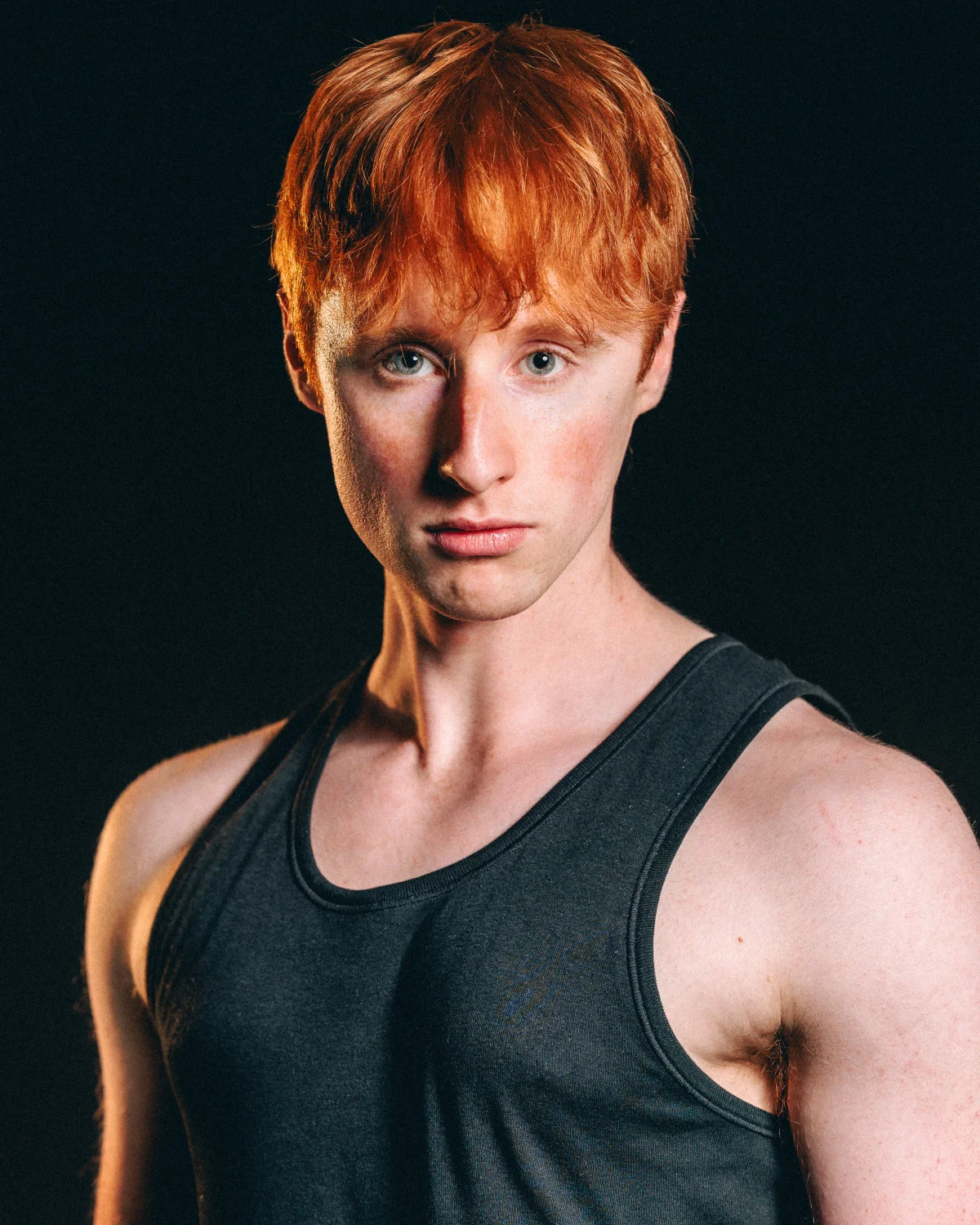 A young man with red hair and blue eyes wearing a grey tank top, posing against a dark background.
