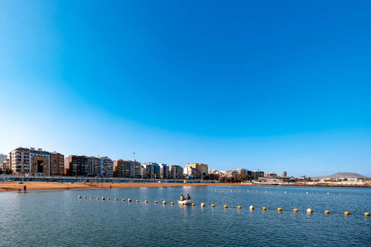 Wide shot of a coastal city skyline bordering a calm blue bay with yellow marker buoys and people on a small boat.