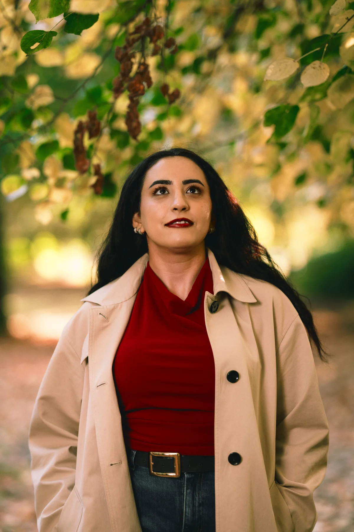 Woman with dark hair in a red top and beige trench coat, looking up amidst autumn leaves.