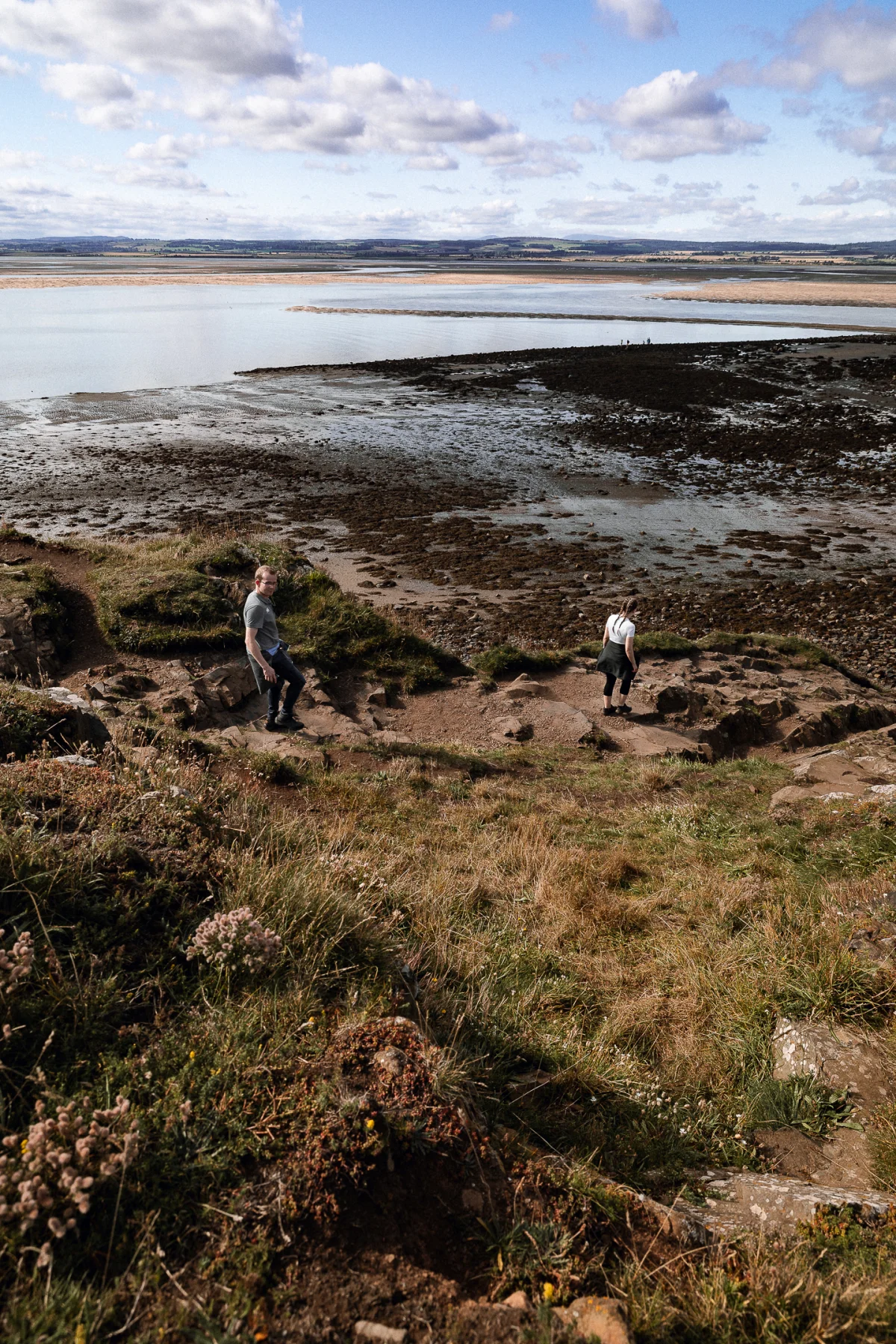 Two people standing on a rocky shoreline overlooking a wide tidal flat under a cloudy sky.
