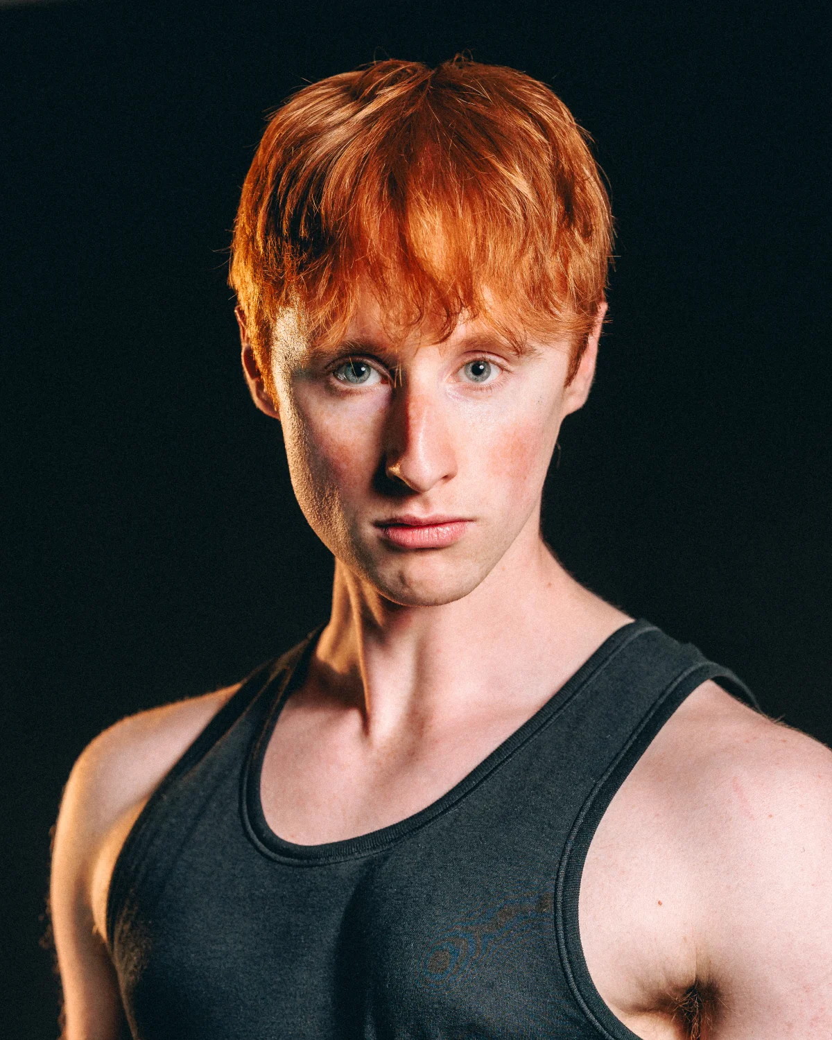 Close-up portrait of a young man with red hair in a grey tank top against a dark background.