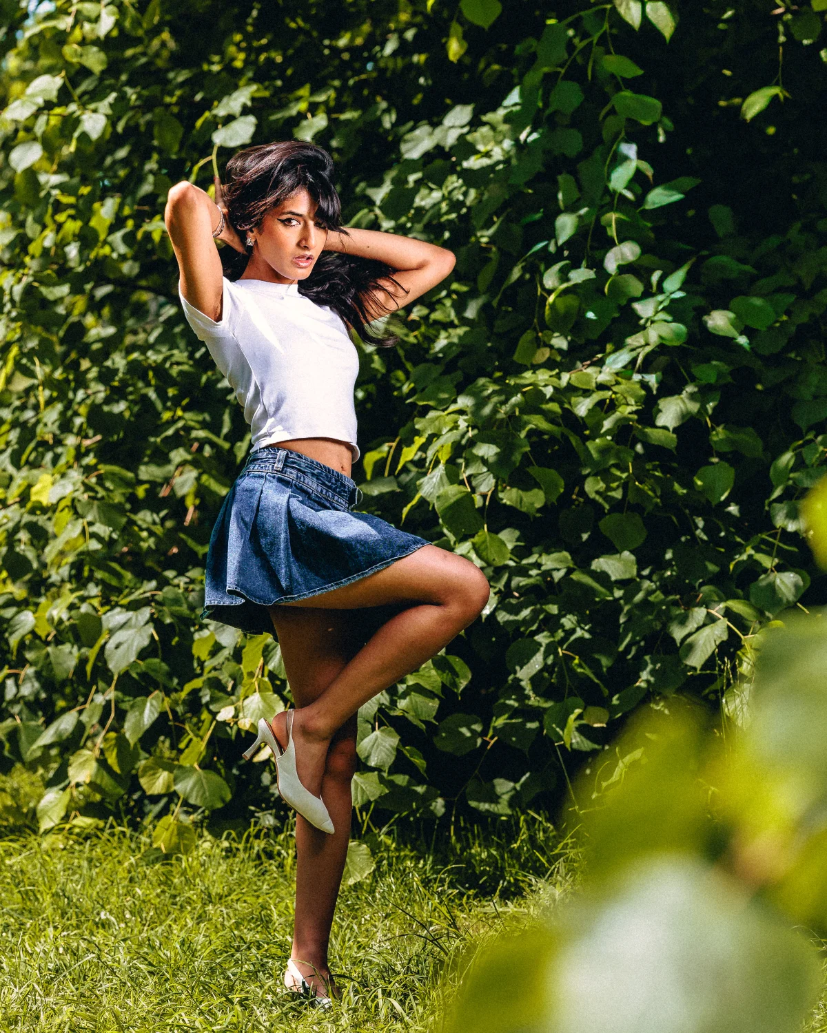 Woman in white crop top and denim mini-skirt posing against a green hedge background.