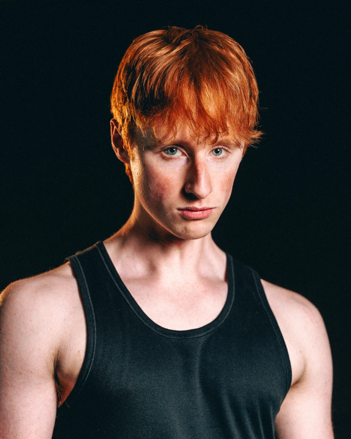Portrait of a young man with red hair in a black tank top against a dark background.