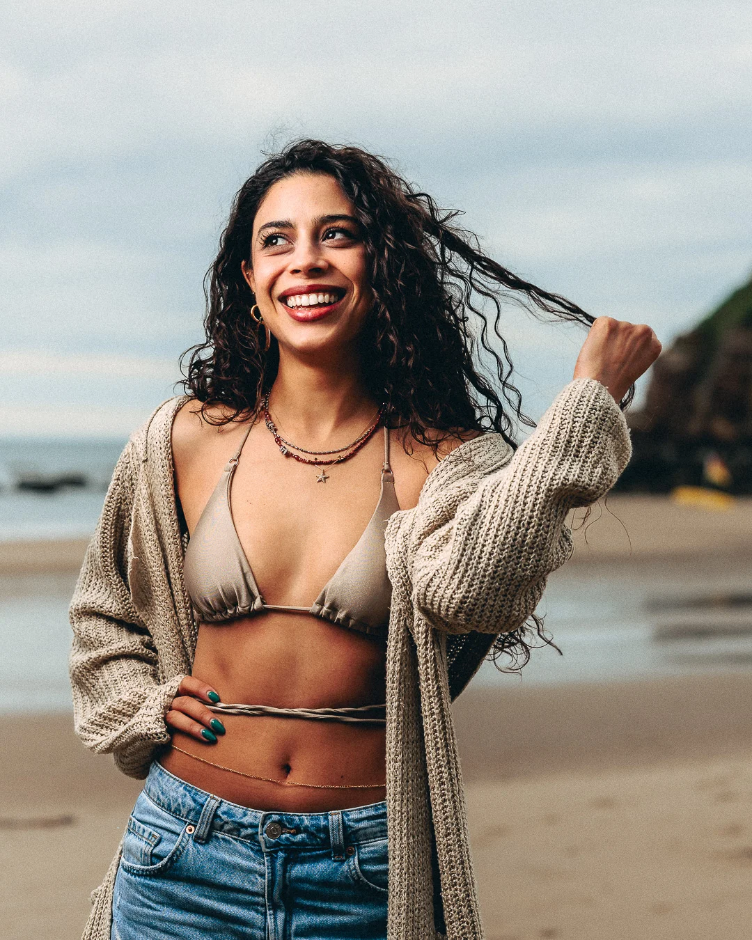 Smiling woman with curly hair posing on a beach in a bikini top, jeans, and an open cardigan.