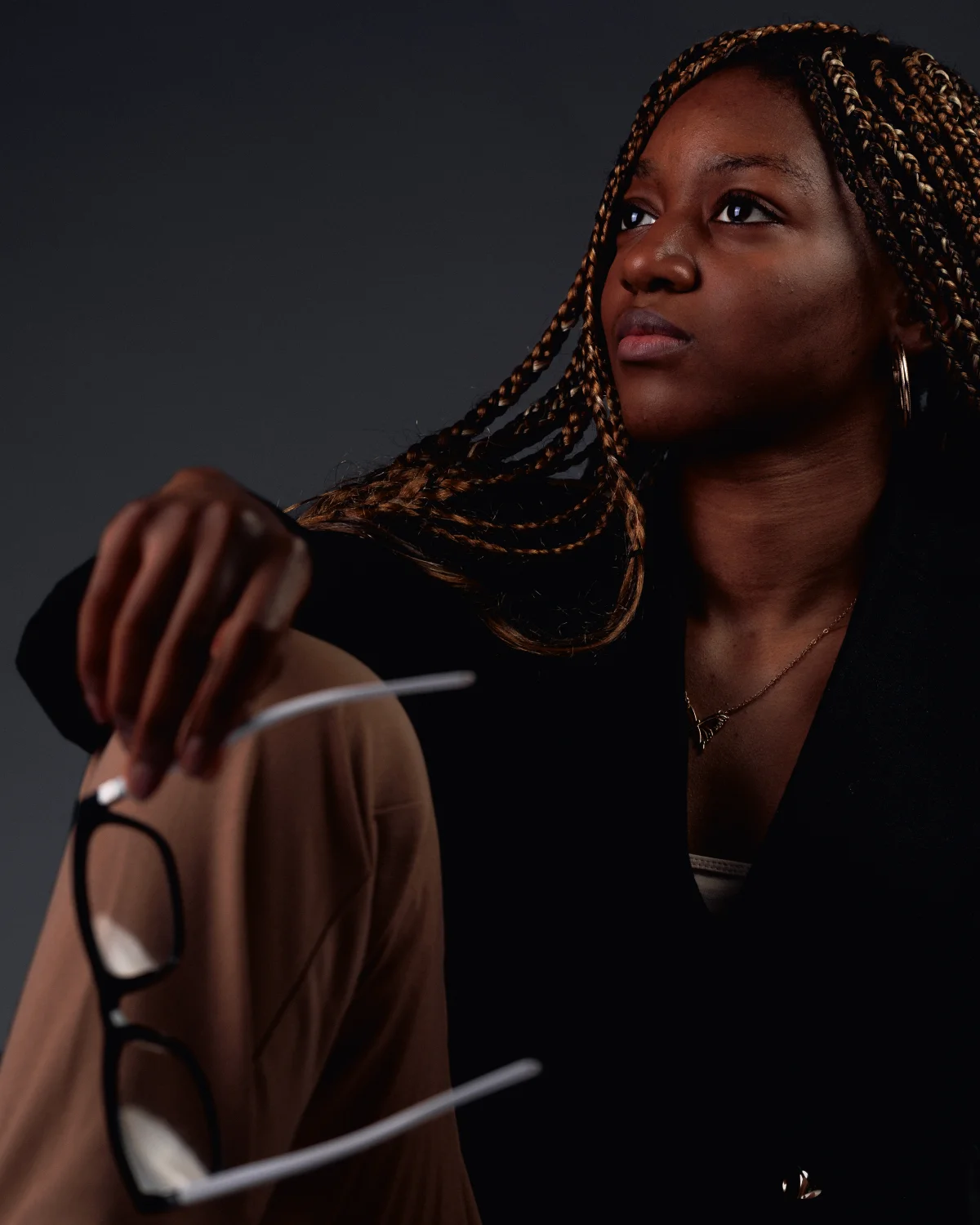 A woman with long braids in a black blazer looks thoughtfully off-camera, holding glasses against a dark background.
