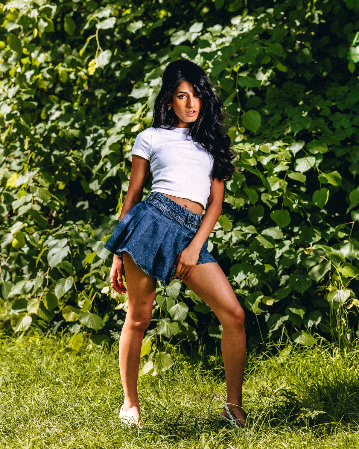 A young woman with long dark hair wearing a white crop top and denim mini skirt posing in front of lush green foliage.