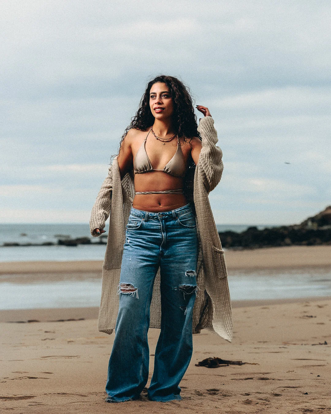 A woman with curly hair posing on a sandy beach in a gold bikini top, long beige cardigan, and ripped blue jeans.