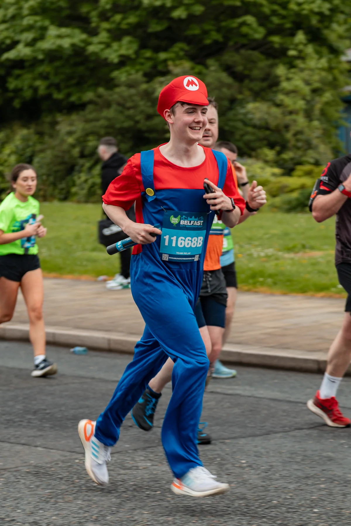 A man dressed as Mario running in a race on a city street with other runners and green trees in the background.
