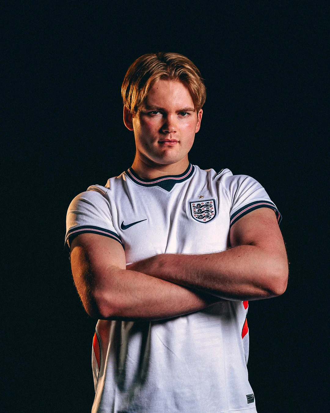 A young man with red hair wearing an England football jersey, arms crossed against a dark black background.