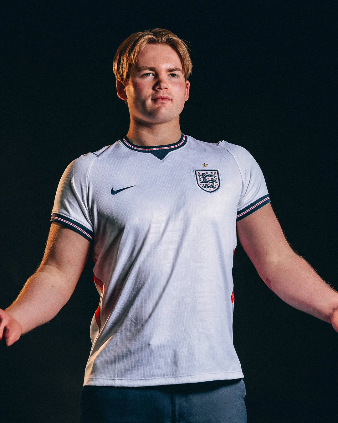 A young man with blond hair wearing an England football jersey against a dark studio background.