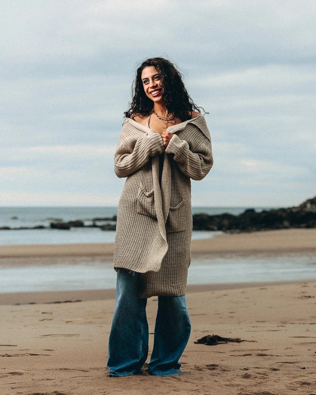Smiling woman with curly hair in a cozy oversized cardigan and flared jeans standing on a sandy beach.
