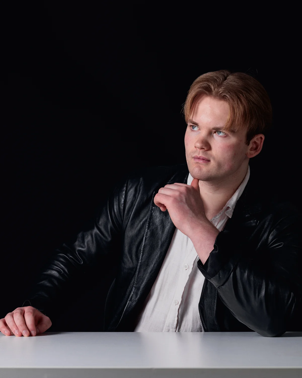 A man with reddish hair in a black leather jacket and white shirt, gazing thoughtfully against a dark background.
