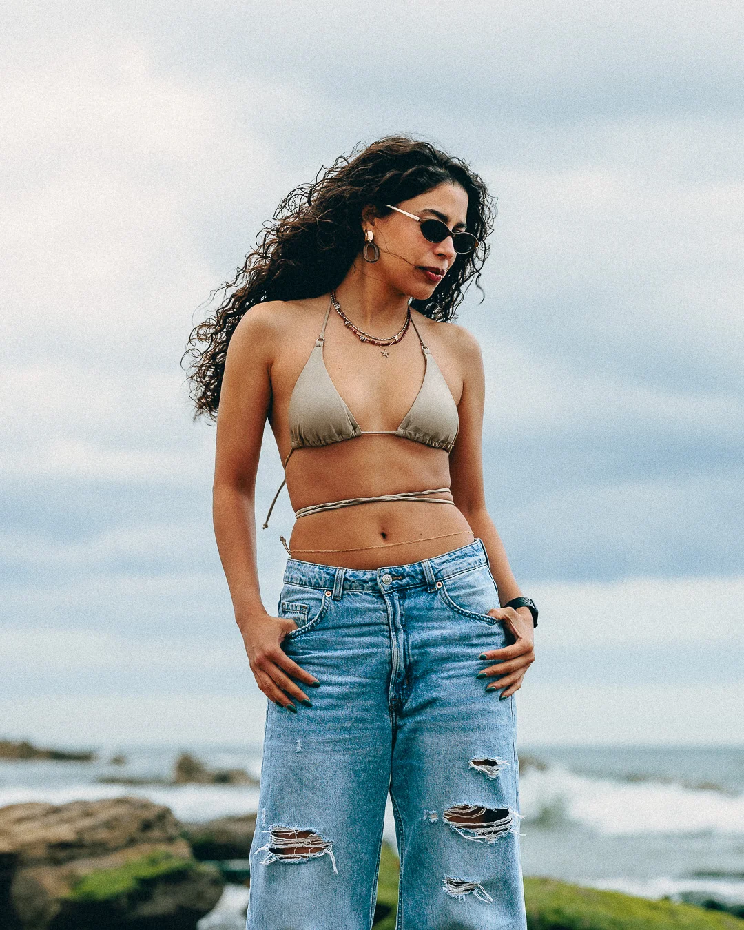 Woman with curly hair in a gold bikini top and ripped blue jeans posing on a beach under an overcast sky.