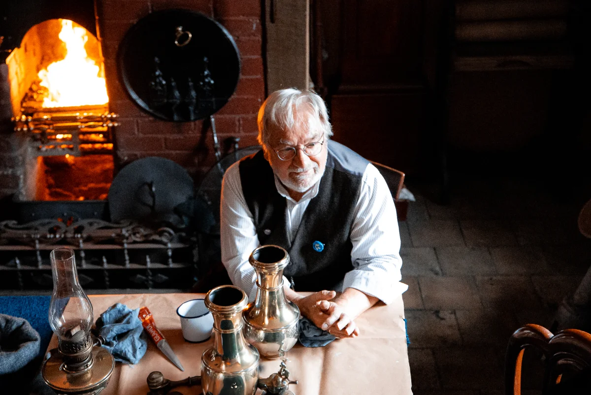 A middle-aged man with white hair and glasses sits at a table with antique copper vessels near a glowing fireplace.