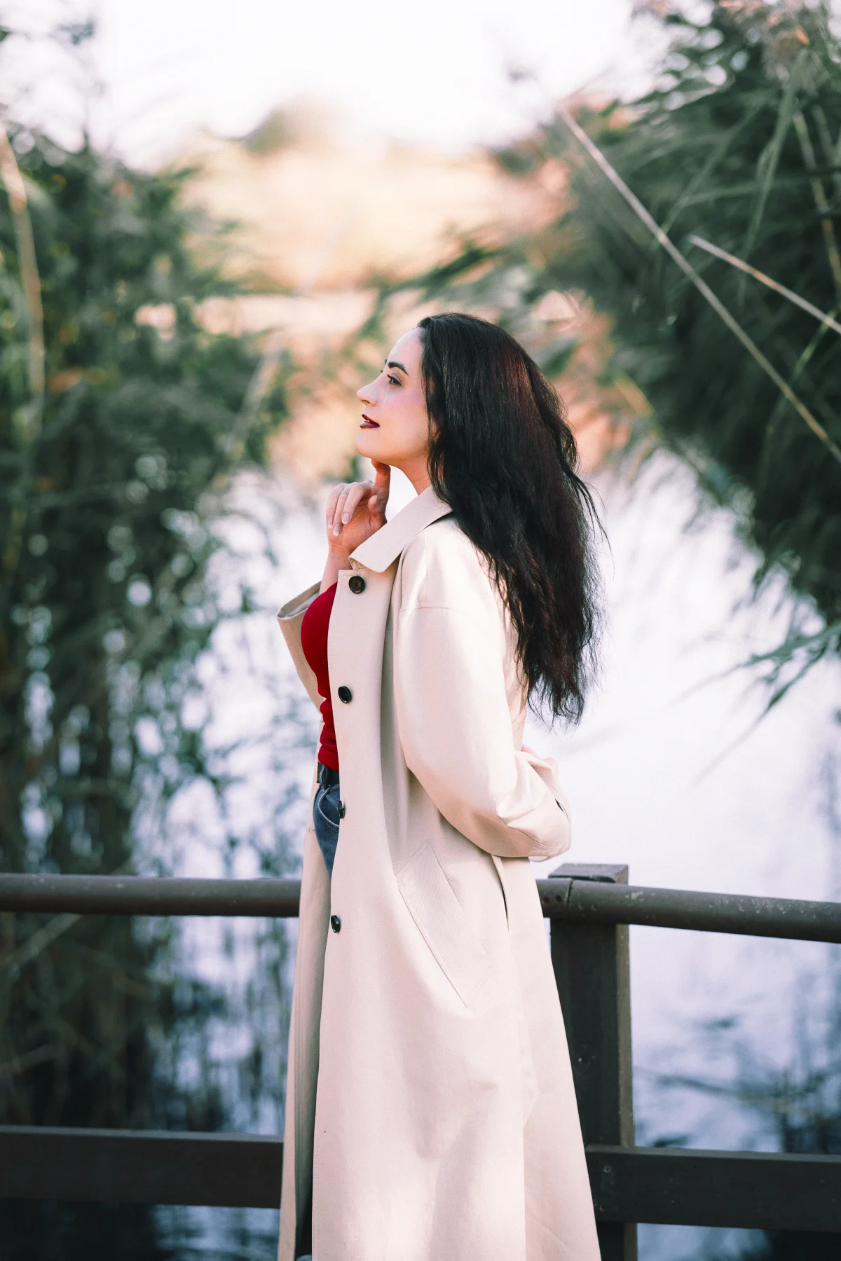 Woman in a beige trench coat posing thoughtfully outdoors with a blurred natural background.