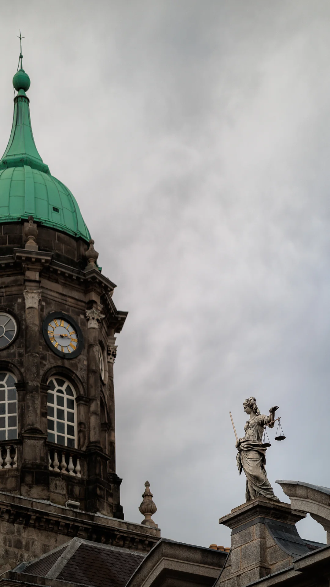 A stone clock tower with a green dome and a statue of Lady Justice under a cloudy sky.
