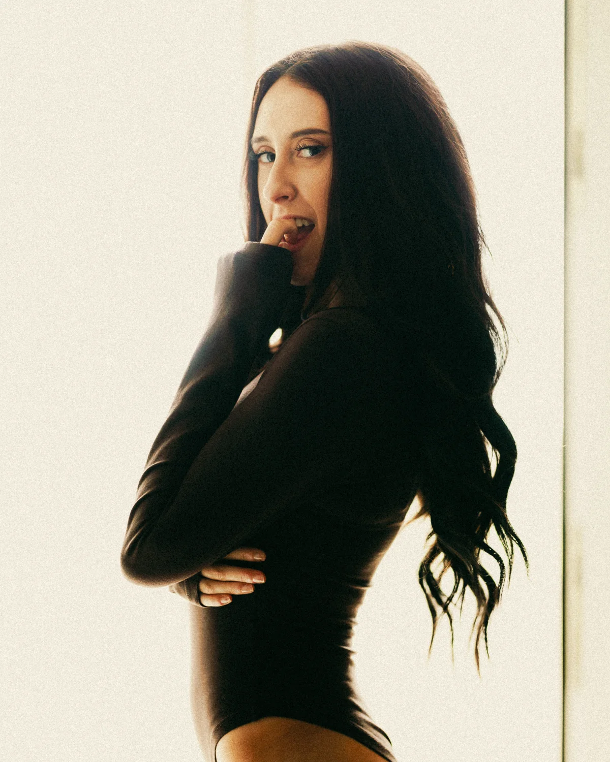 A woman with long dark hair in a black bodysuit biting her lip, posing against a bright neutral background.