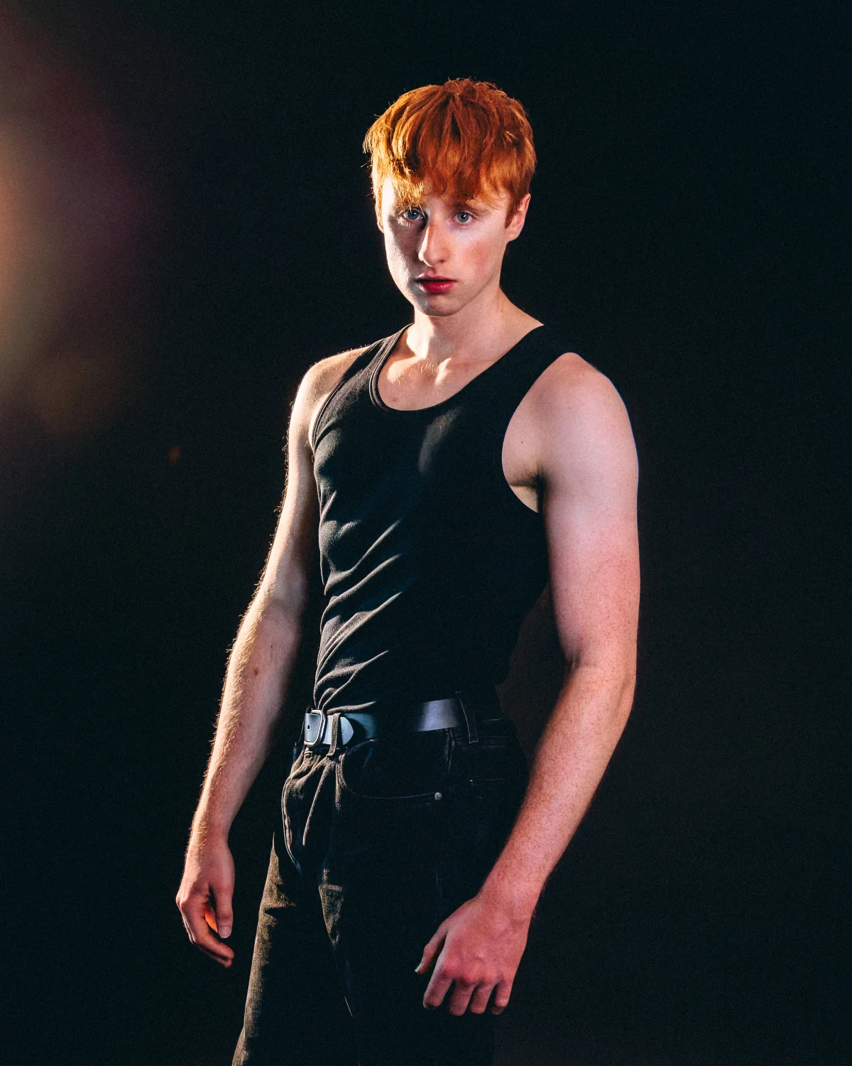 A young man with red hair in a black tank top posing against a dark studio background.