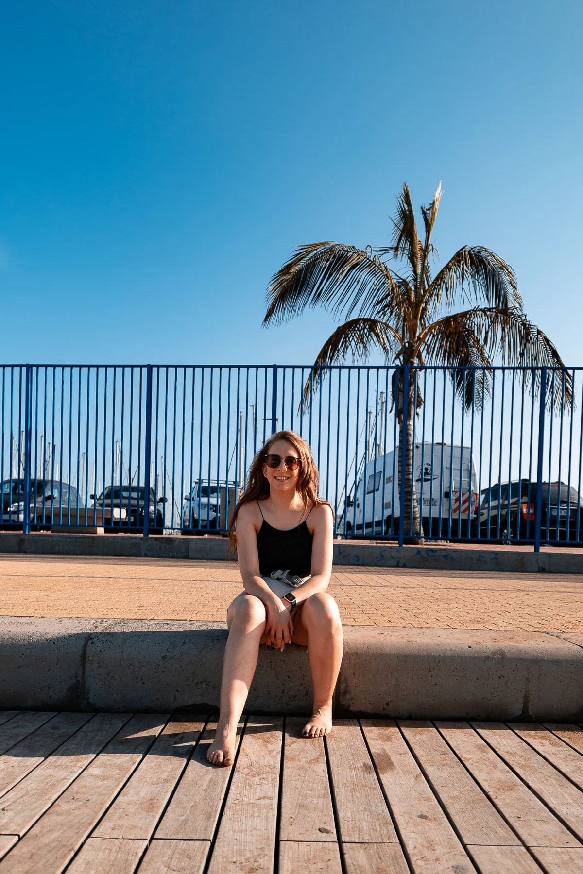 A smiling woman wearing sunglasses sitting on a concrete ledge outdoors with a palm tree and blue fence behind her.