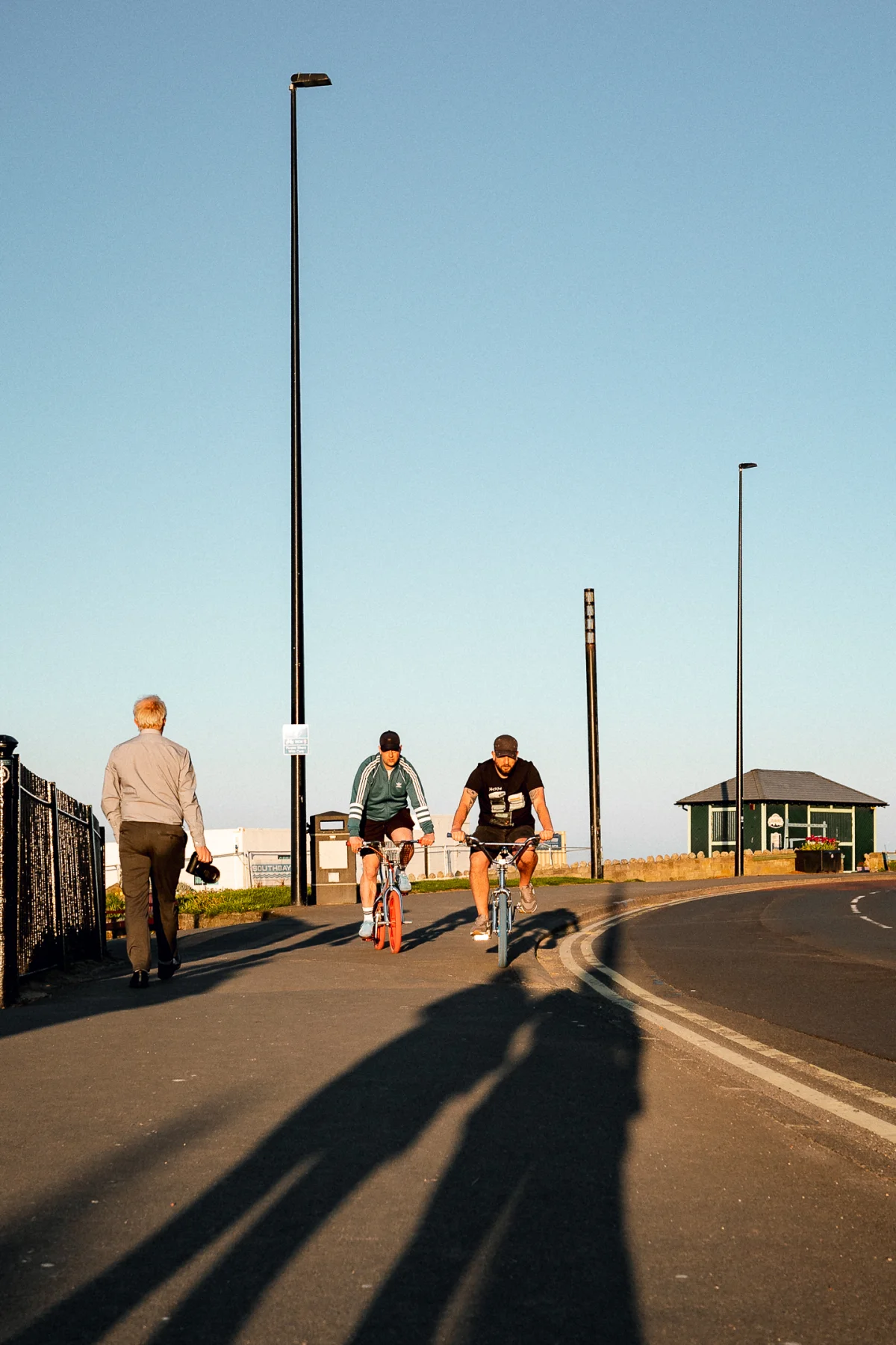 Two cyclists ride toward the viewer on a sunny coastal path, with a man walking away in the foreground.