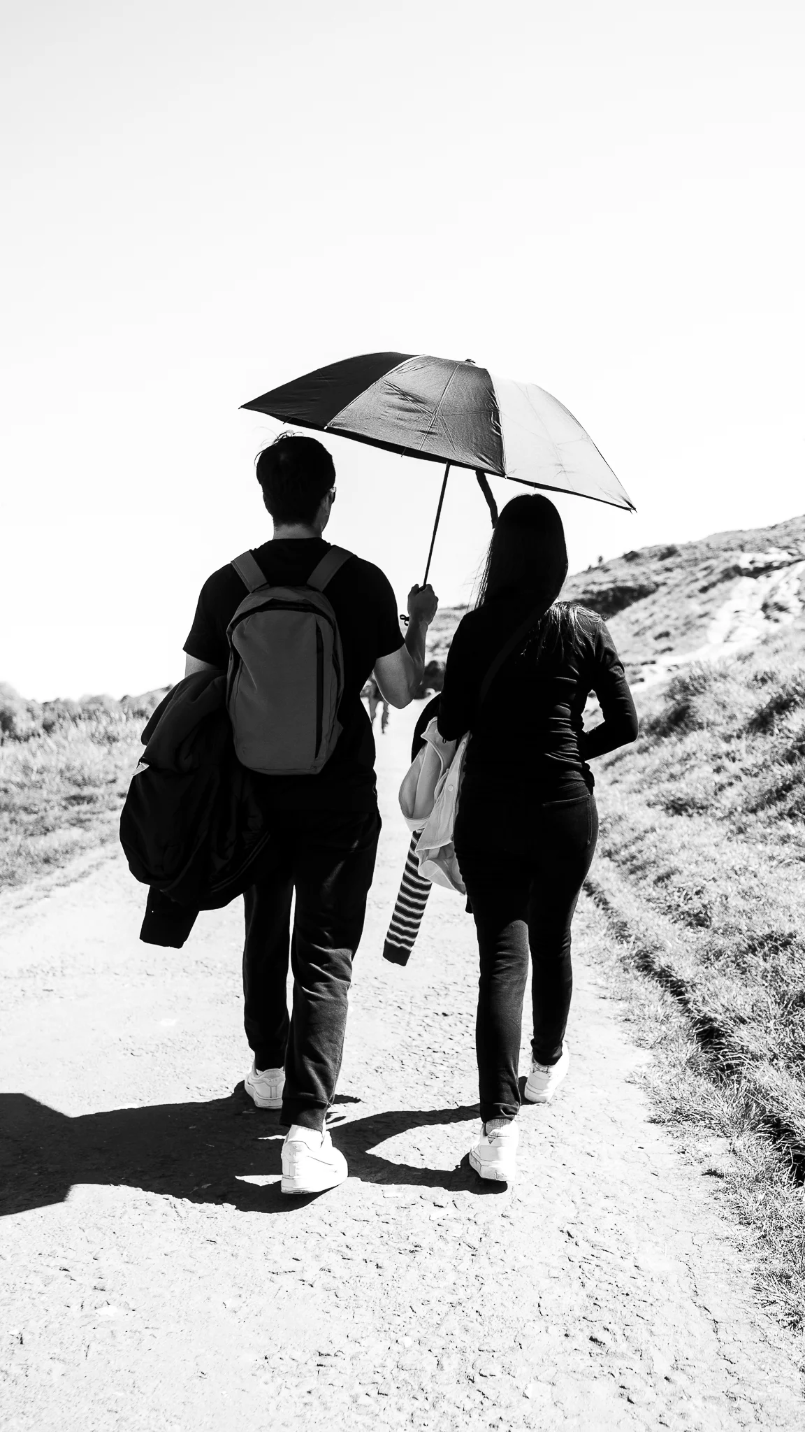 Black and white photo of a couple walking away on a path under an umbrella in a hilly landscape.