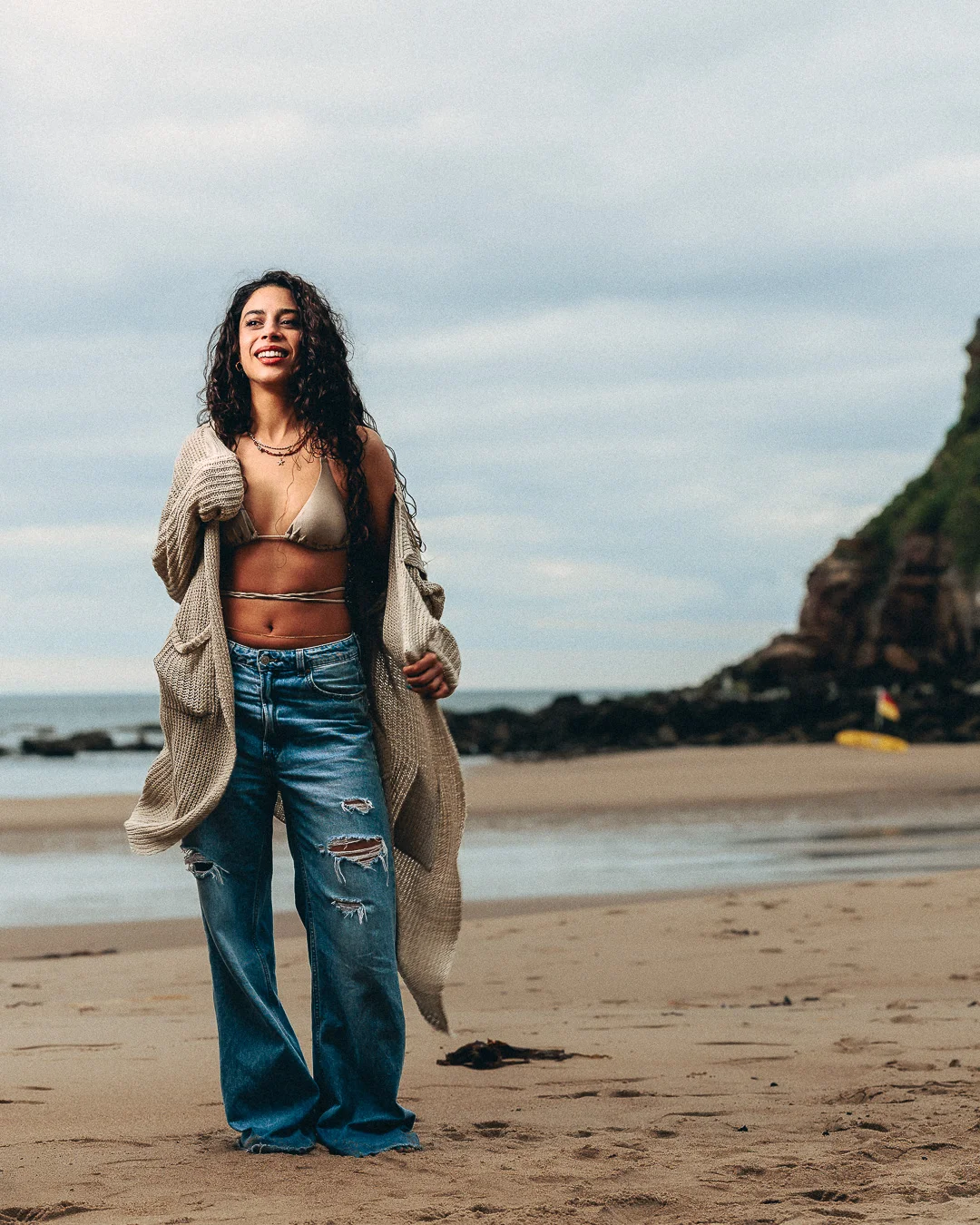 A smiling woman with curly hair on a sandy beach, wearing a bikini top, oversized cardigan, and ripped wide-leg jeans.
