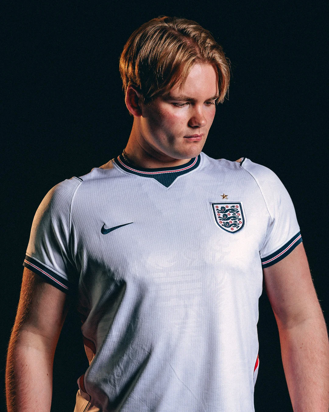 A moody studio portrait of a young man wearing an England football jersey against a dark background.