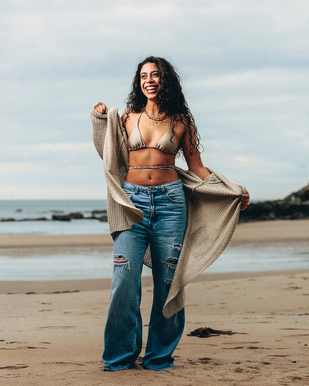 Smiling woman with curly hair in a bikini top and flared jeans on a sandy beach.