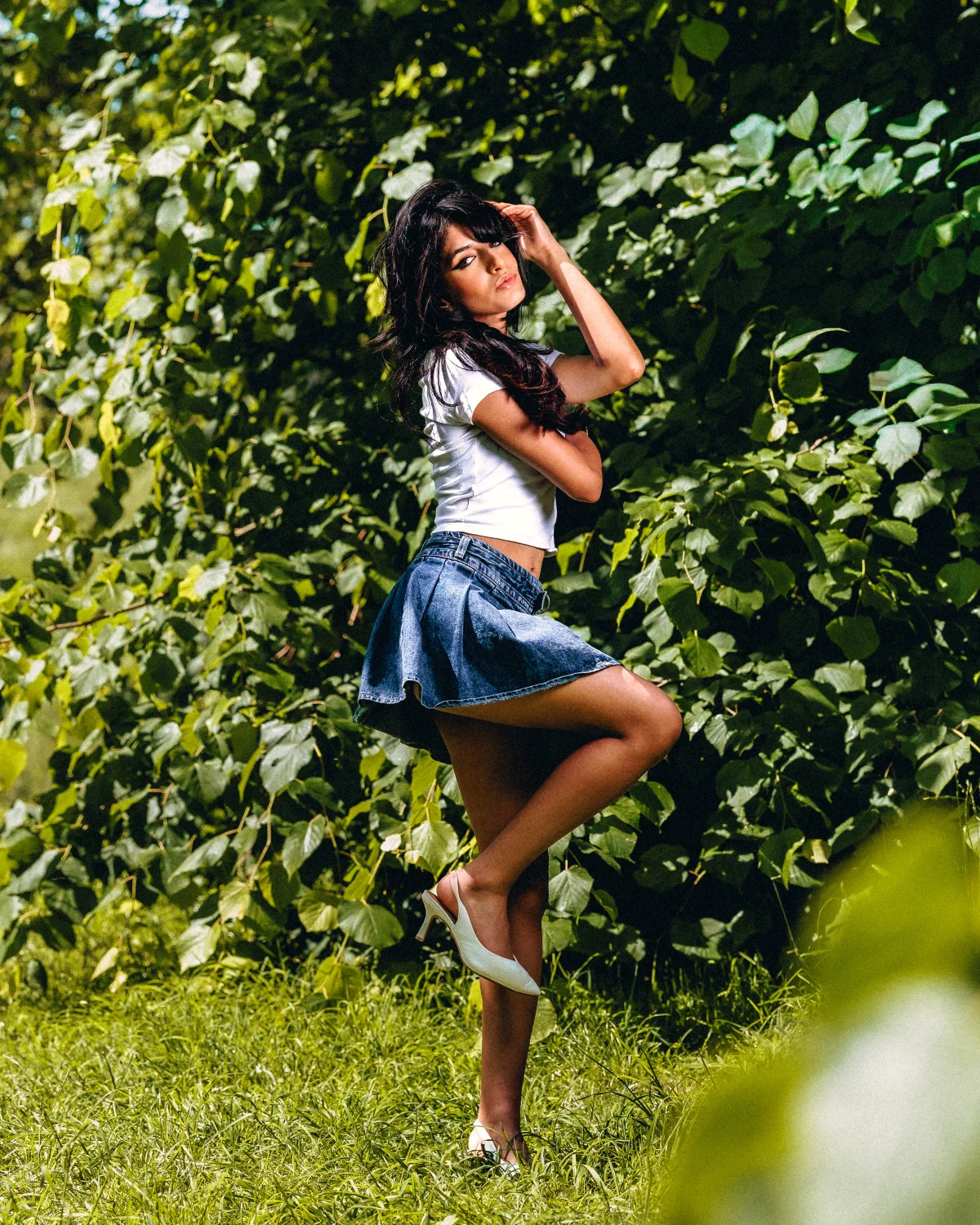 A woman in a white t-shirt and denim mini skirt posing against a lush green foliage background.