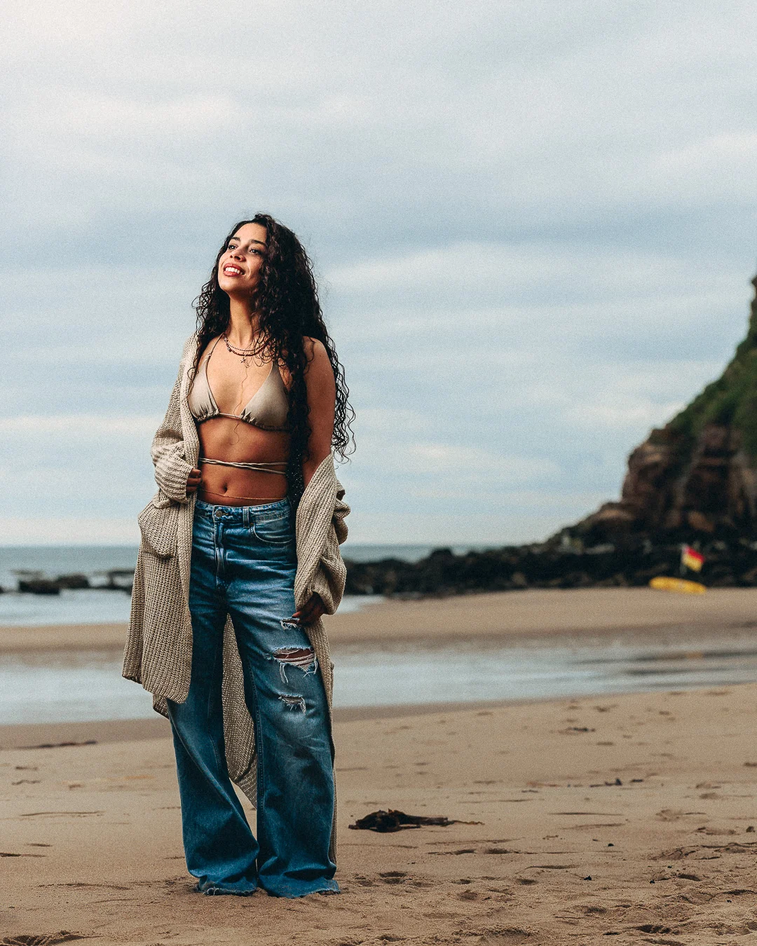 A woman with long curly hair posing on a sandy beach, wearing a bikini top, oversized beige cardigan, and wide-leg jeans.