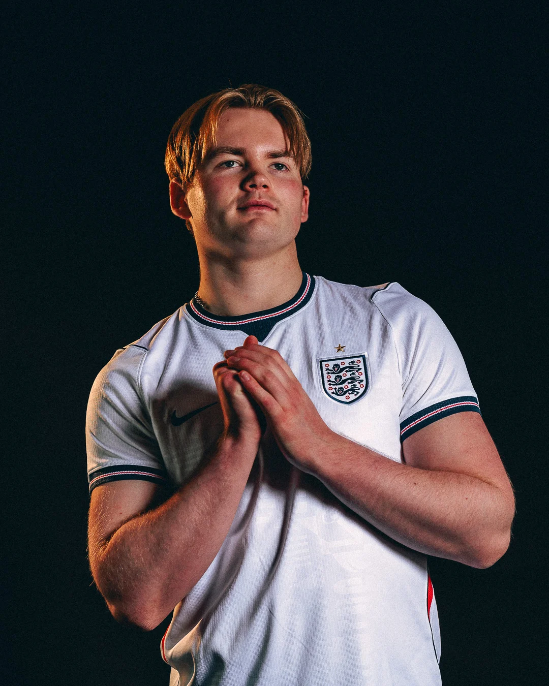 Dramatic studio portrait of a young man in an England football jersey against a black background.