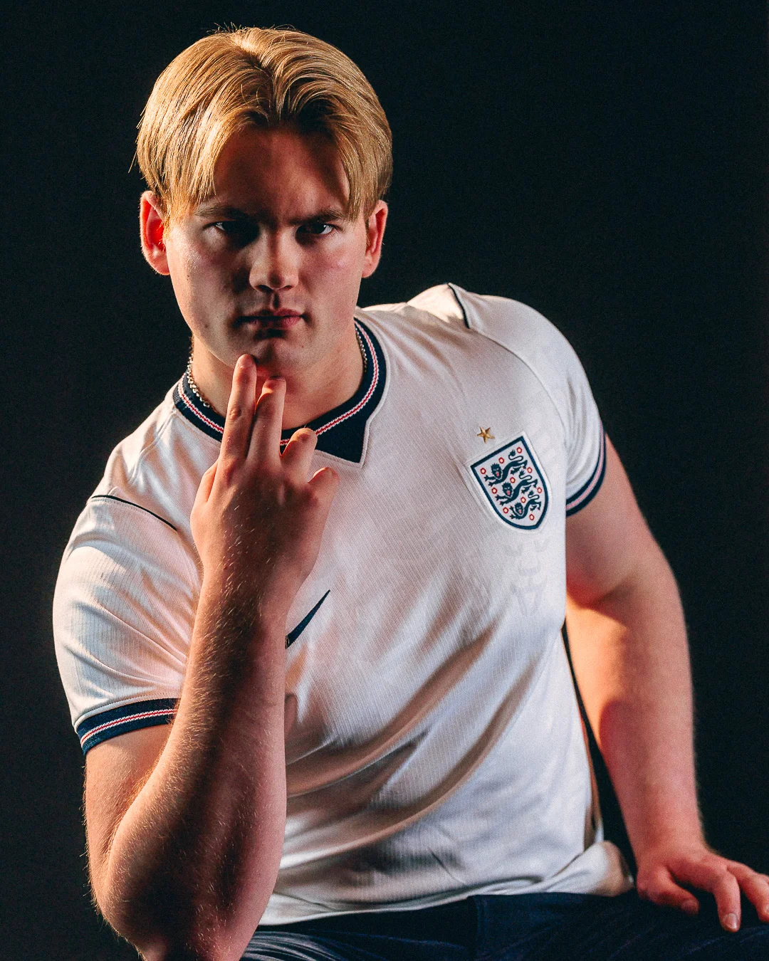 A young man with blonde hair in a white England football jersey posing thoughtfully against a dark background.