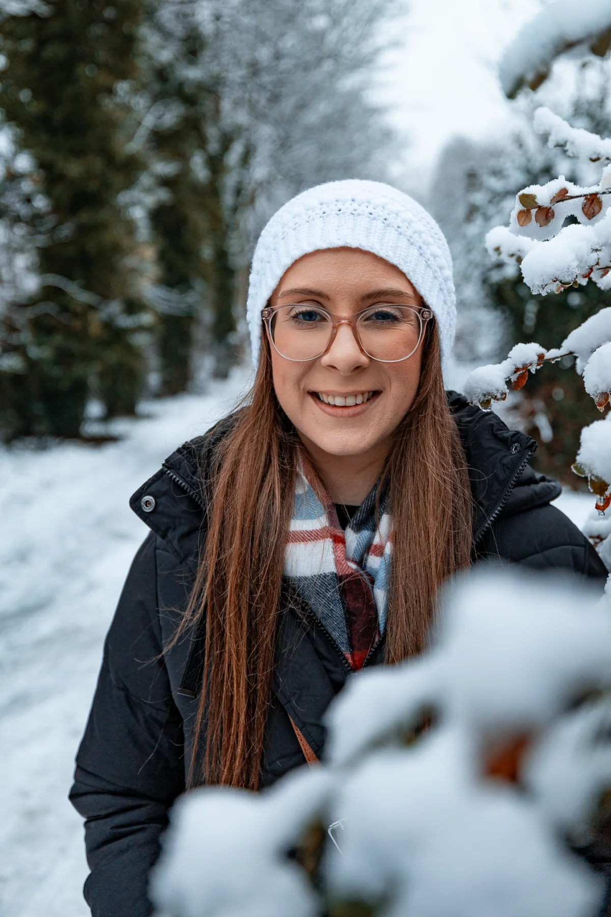 A smiling woman wearing a white beanie and glasses in a snowy winter forest.