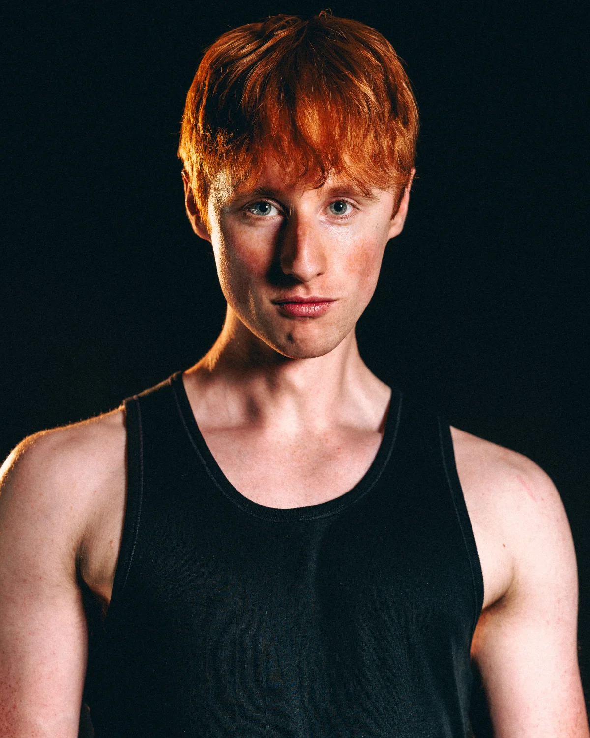 A close-up studio portrait of a young man with red hair wearing a black tank top against a dark background.