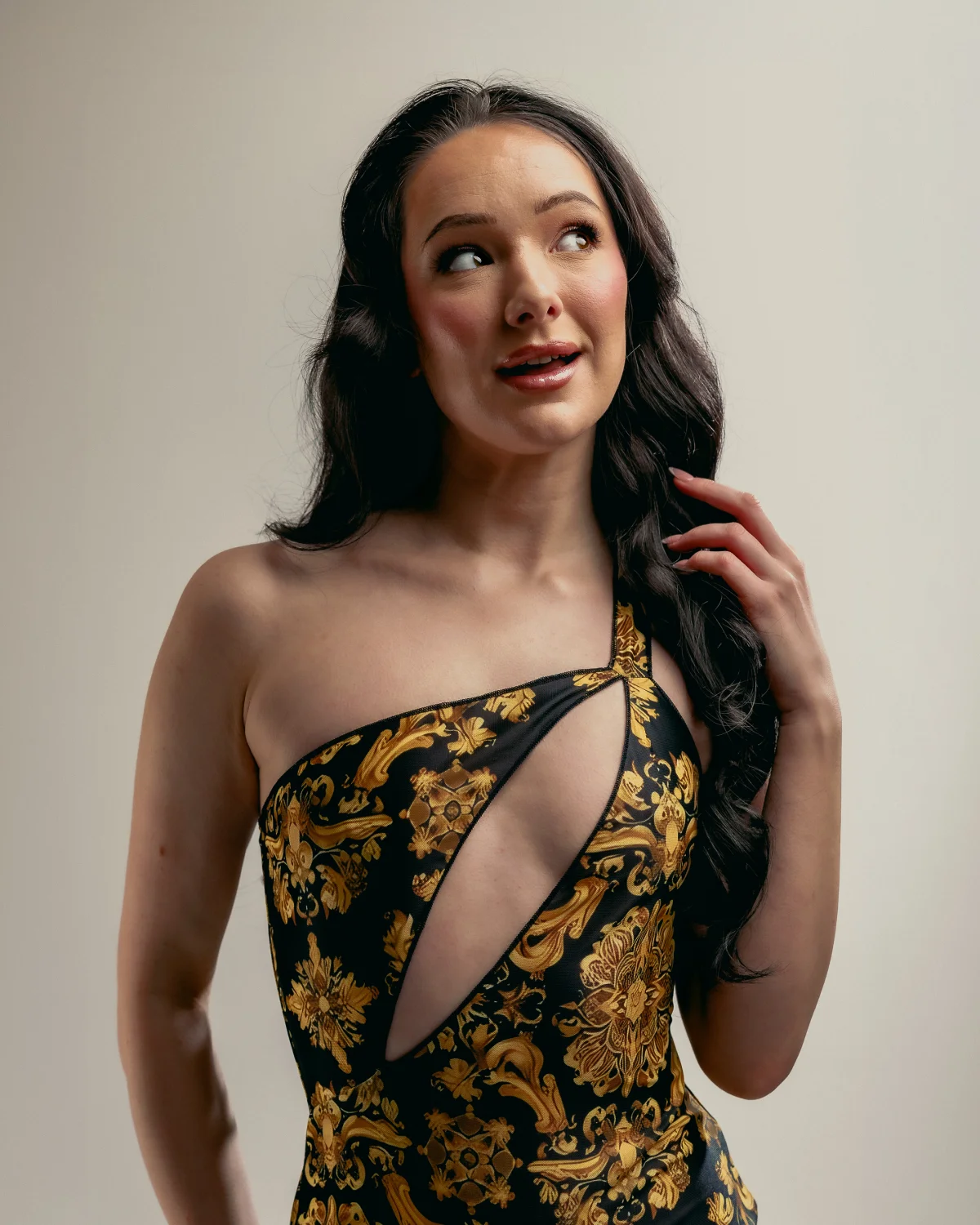 Woman with long dark hair in a gold and black patterned one-shoulder dress posing against a neutral background.