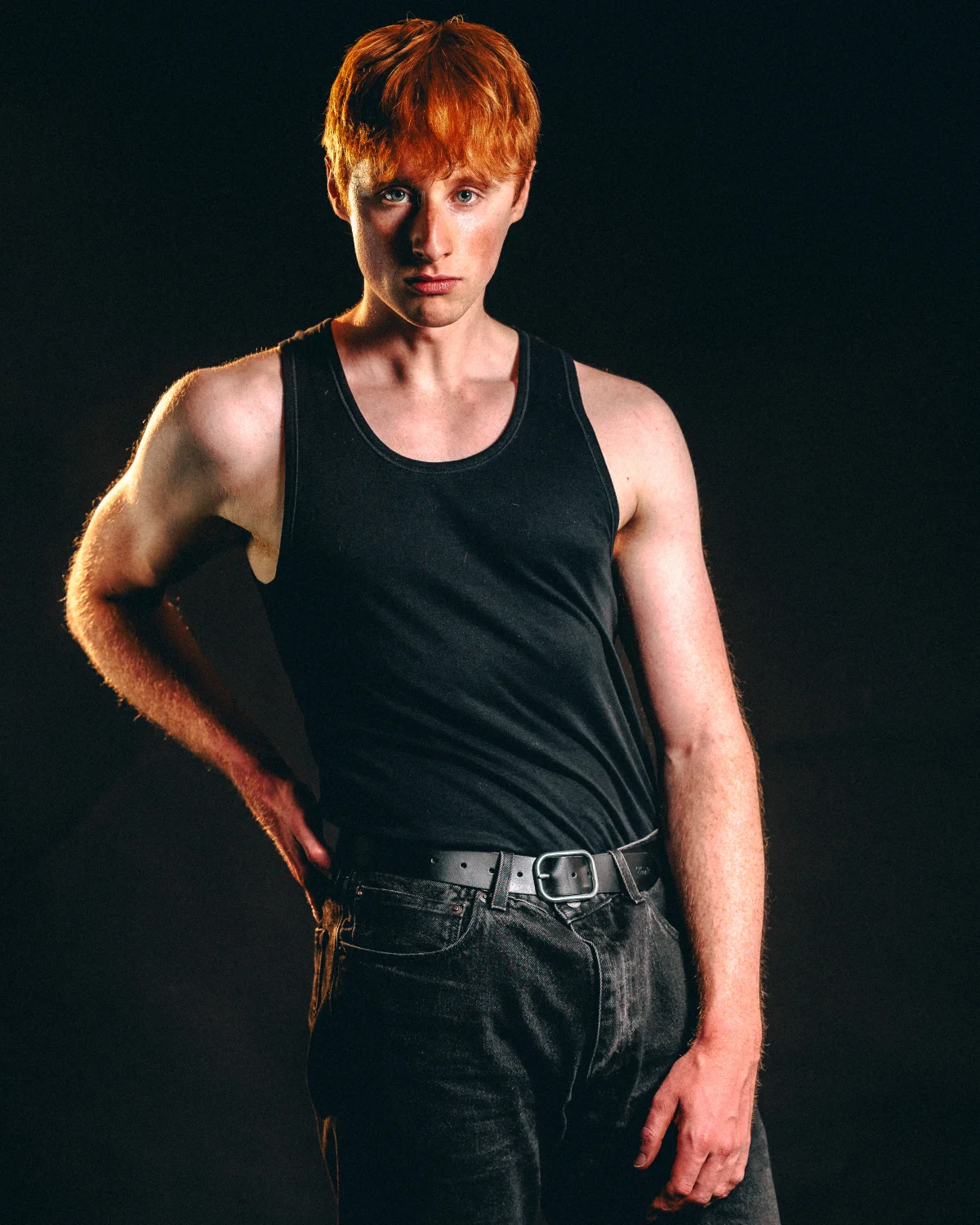 Studio portrait of a red-haired man in a black tank top and dark jeans against a solid black background.