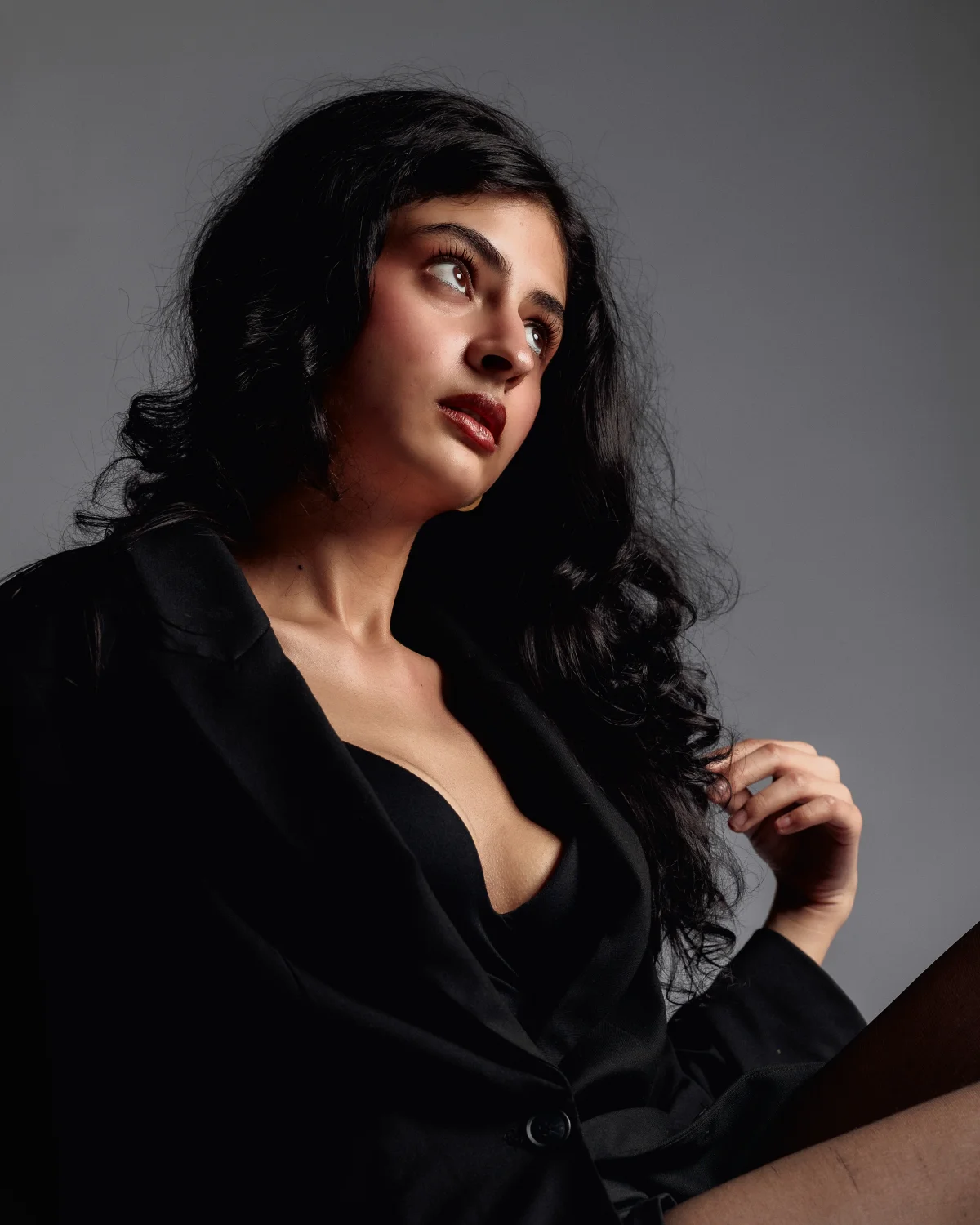 A woman with long dark wavy hair and bold red lipstick wearing a black blazer against a grey studio background.