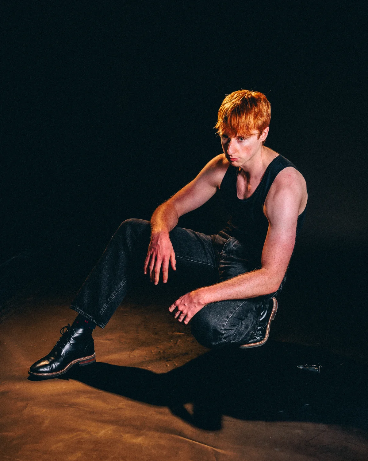 A young man with red hair in a black tank top and jeans sitting on a dark floor under moody studio lighting.
