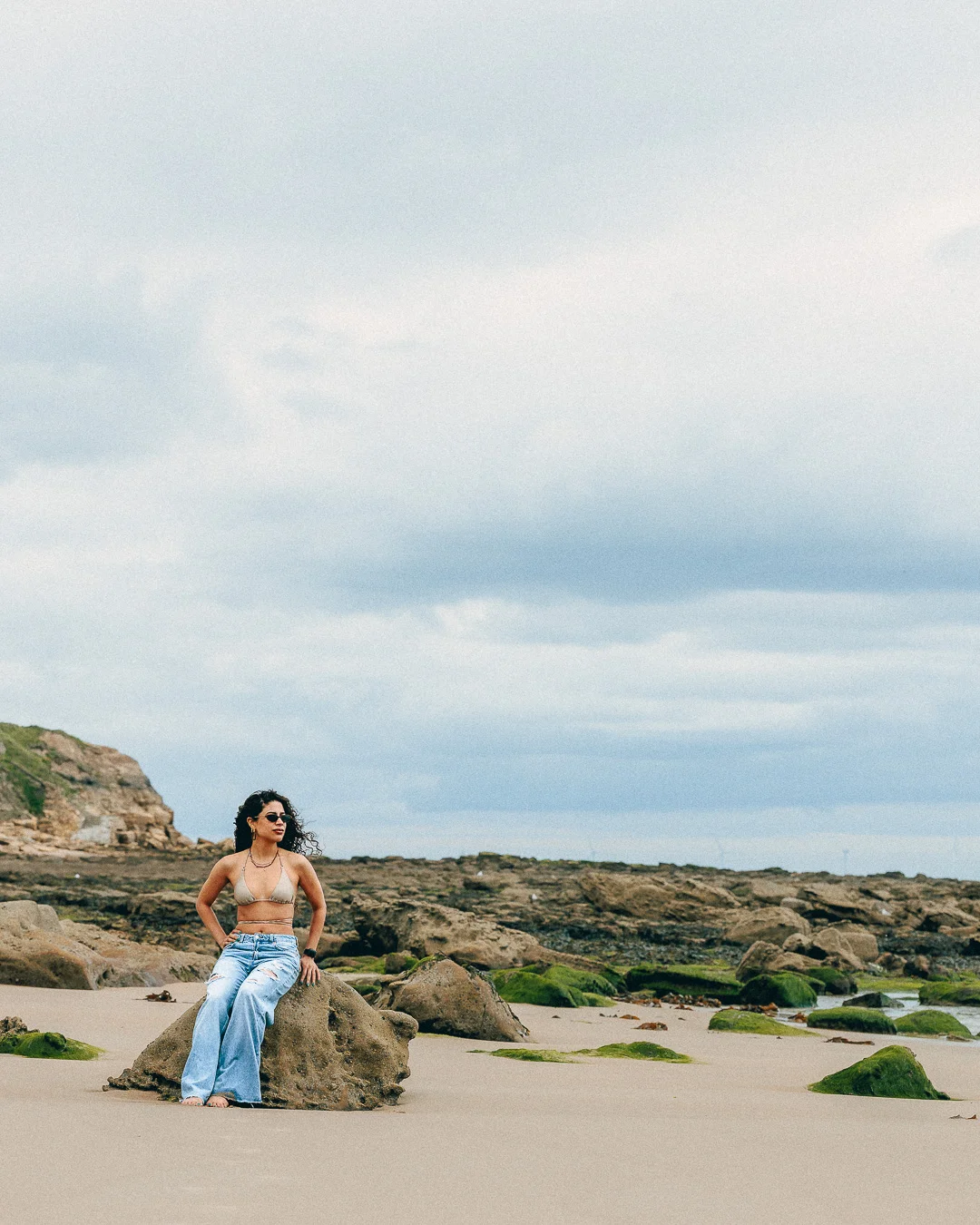 A woman in a bikini top and light blue jeans sitting on a rock at a rocky beach under a cloudy sky.