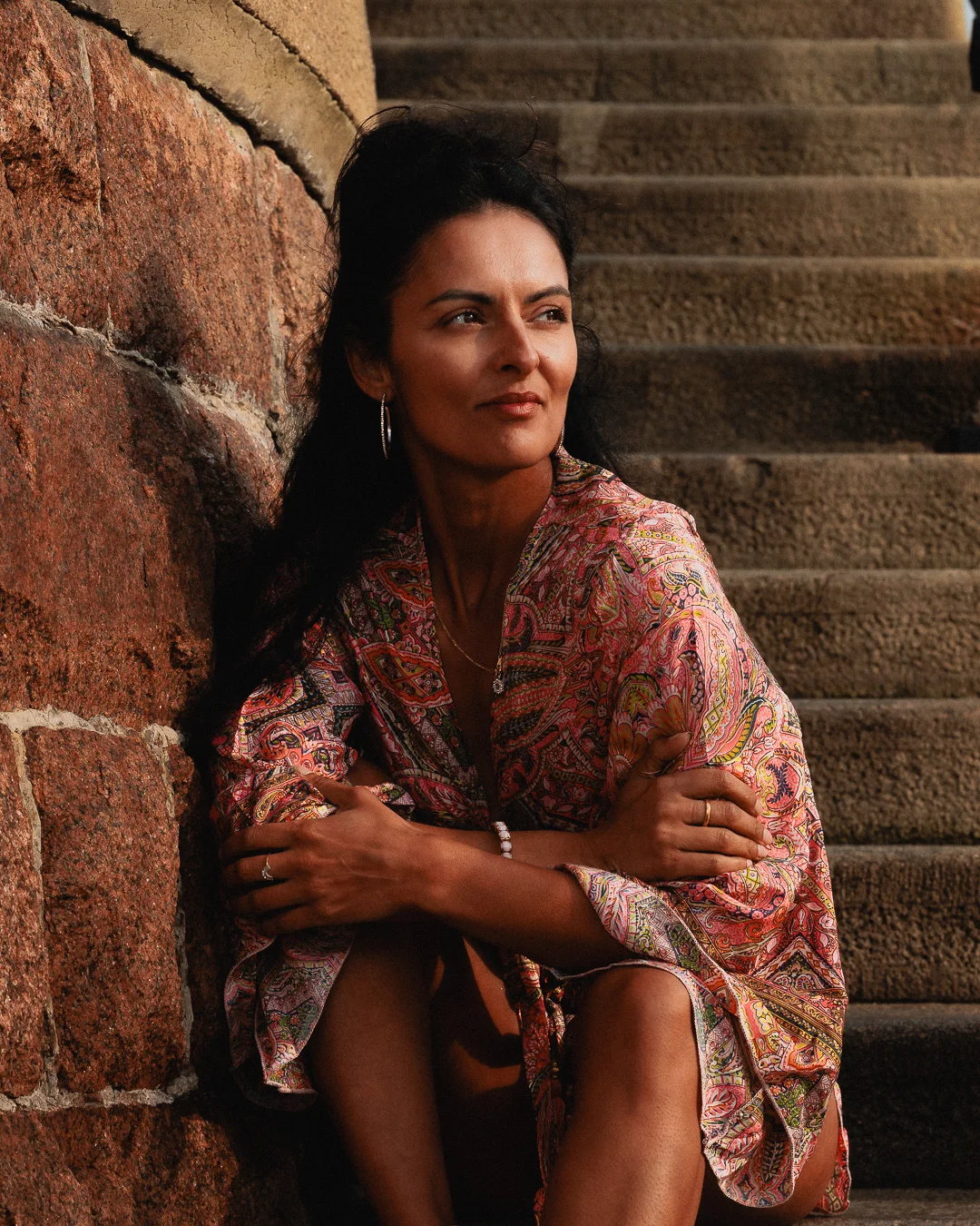 Woman in a colorful paisley dress sitting on stone steps against a textured wall in warm natural light.