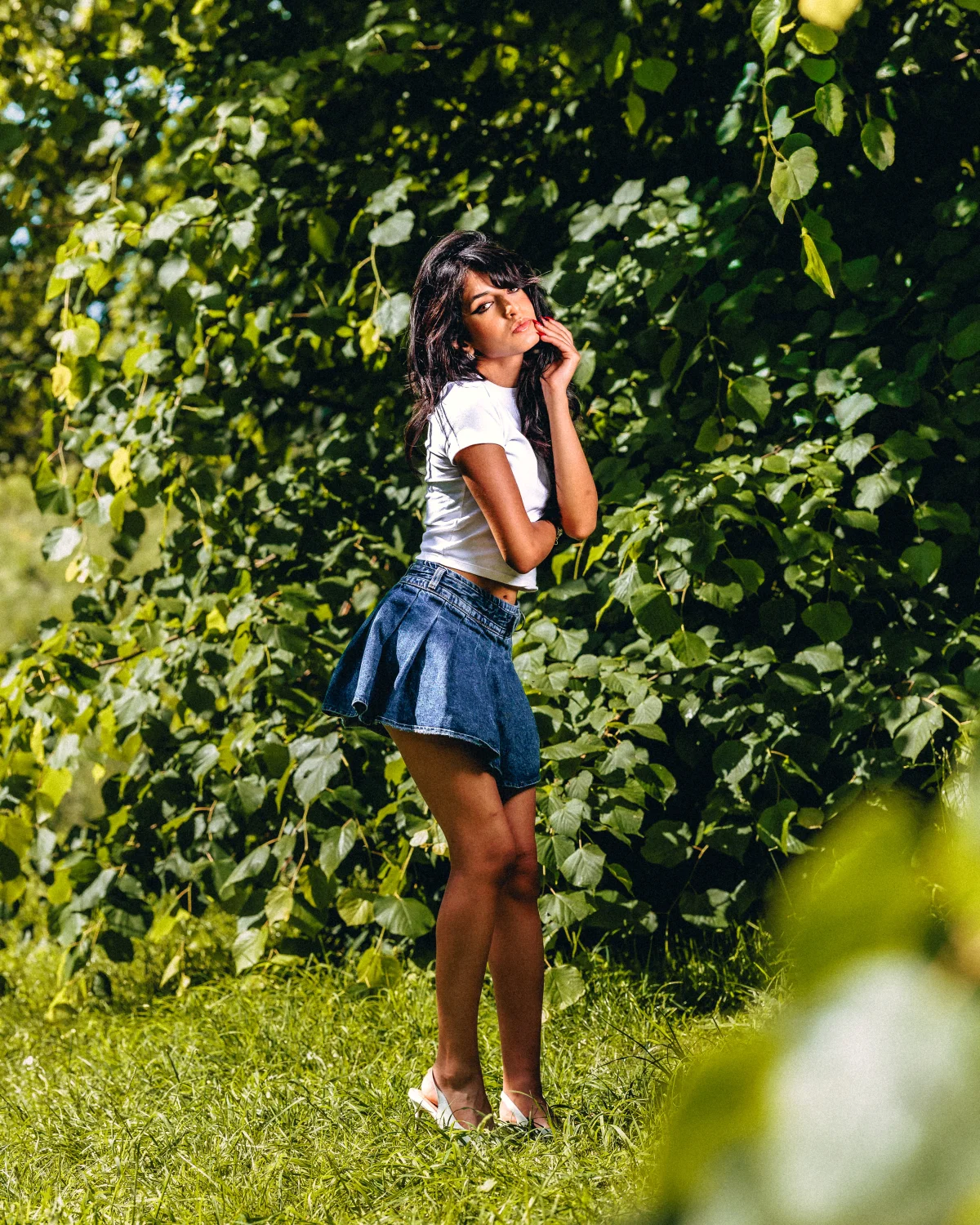 A woman in a white t-shirt and short denim skirt posing against a lush green hedge.