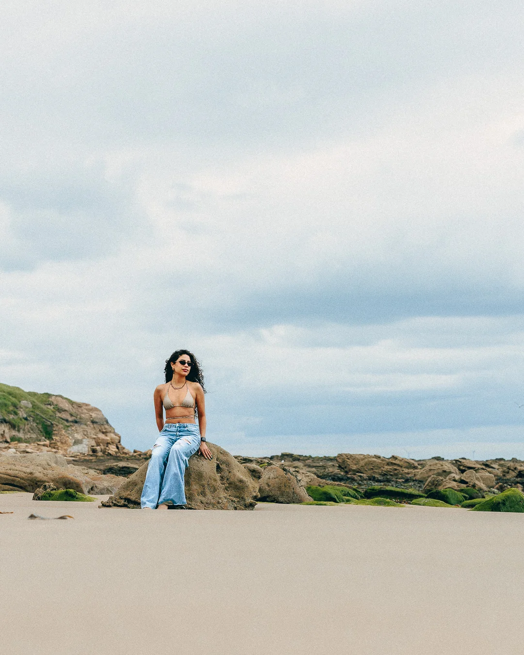 Woman in a bikini top and light blue jeans posing on a rocky beach under an overcast sky.
