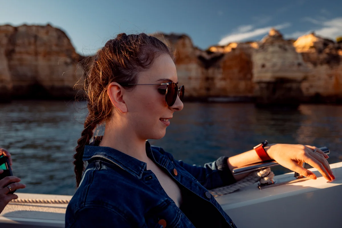 A woman in a denim jacket and sunglasses on a boat during golden hour with coastal cliffs in the background.