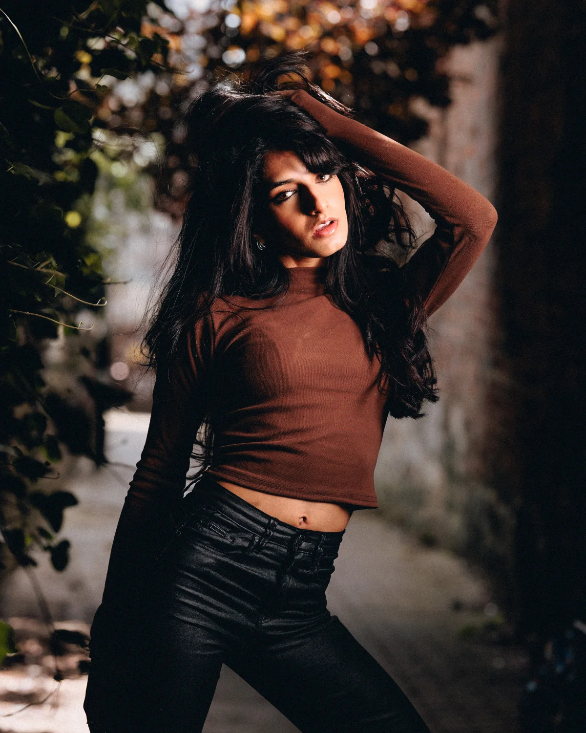 A young woman with long dark hair posing in a brown crop top and black leather pants in an outdoor setting.
