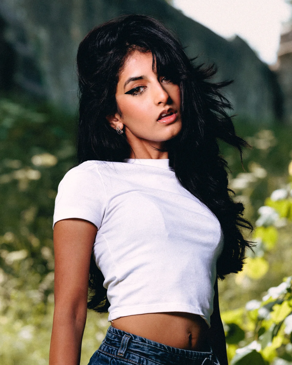 A young woman with long dark hair in a white crop top and blue jeans posing outdoors in natural light.