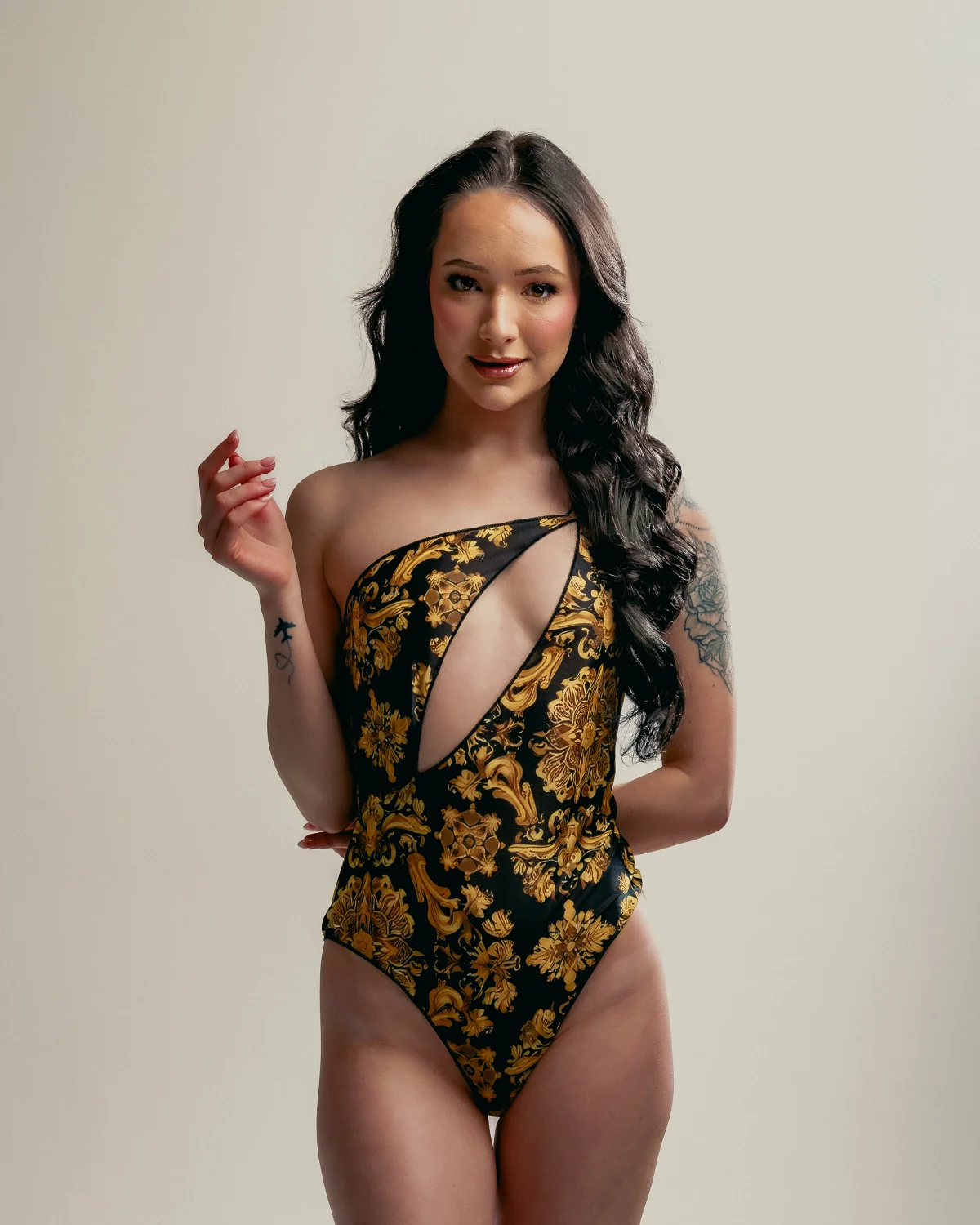 A woman with long dark wavy hair posing in a black and gold patterned one-shoulder bodysuit against a neutral studio background.