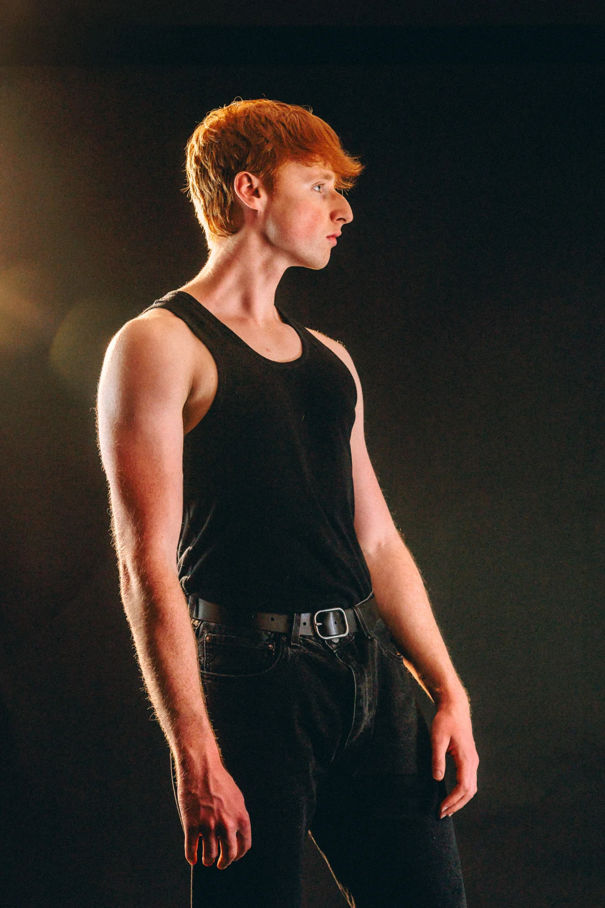 A young man with red hair in a black tank top and jeans posing against a dark background with moody lighting.