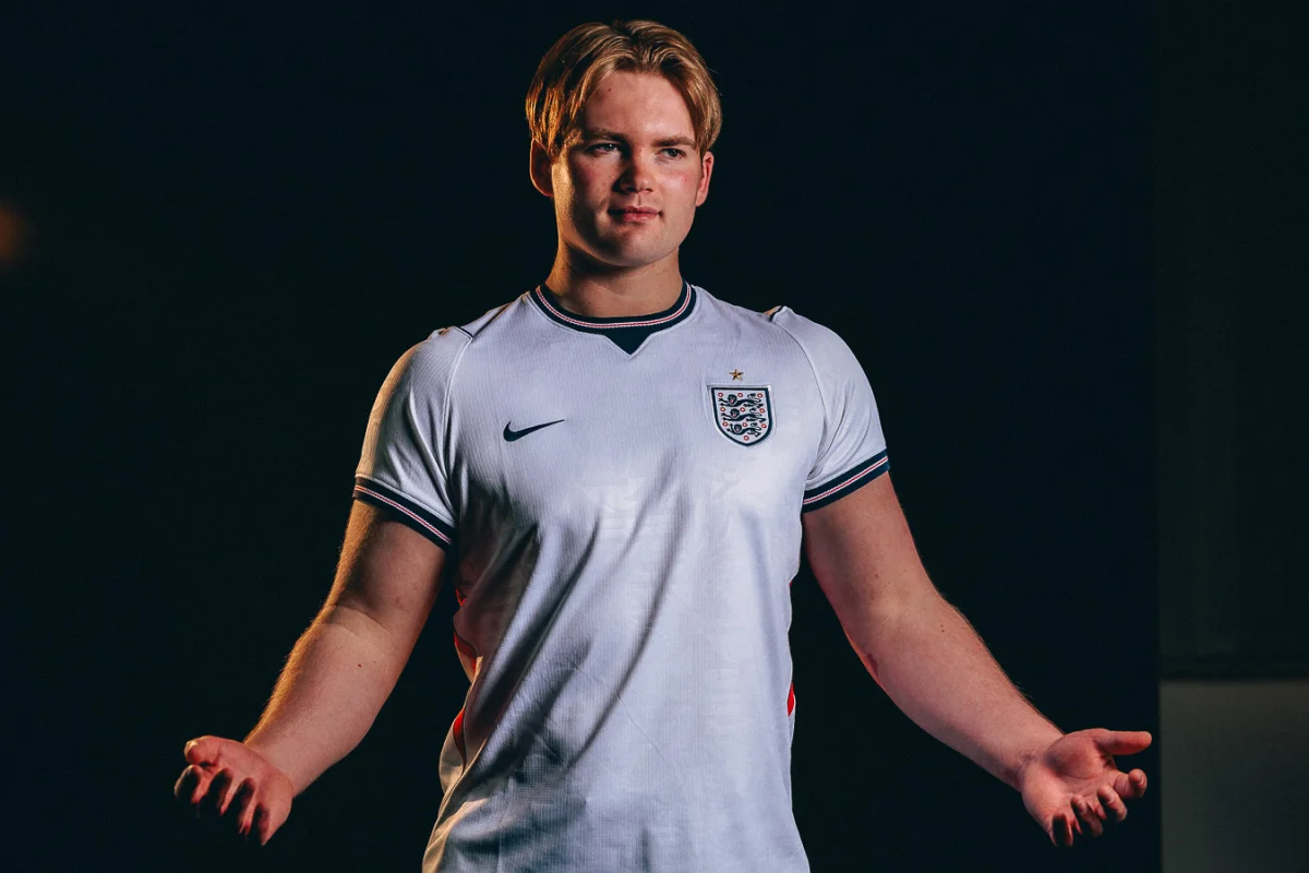 A young man with blond hair wearing an England football jersey against a dark background with dramatic lighting.